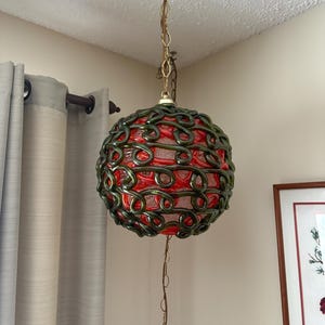 May include: A vintage pendant light. The red glass globe is encased in a green, swirling metal cage. It hangs from a gold chain attached to the ceiling. A unique home decor item. The light fixture is a decorative item.