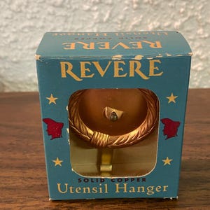 Op de afbeelding: Een blauwe doos met een gouden Revere Utensil Hanger. De hanger is gemaakt van massief koper en heeft een kransontwerp. De doos is versierd met sterren en een rood-witte illustratie van een mensenhoofd aan elke kant.