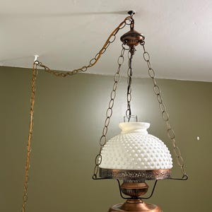 May include: A vintage hanging light fitting featuring a white hobnail glass shade and copper-coloured metal detailing. The light is suspended from the ceiling by a chain and a cord. The design is classic and retro.