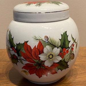 May include: White ceramic jar with a lid, decorated with a red poinsettia, green holly leaves, and white flowers. The jar has a gold trim around the lid.