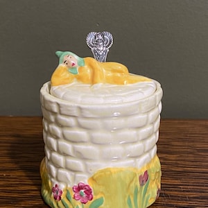 Könnte beinhalten: Ein Keramik-Honigtopf in Form eines Ziegelbrunnens mit einer gelben und grünen Figur, die obenauf liegt. Die Figur trägt einen grünen Hut und gelbe Kleidung. Der Topf hat einen silbernen Löffel mit einem dekorativen Griff.