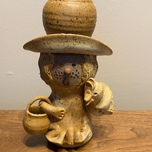 May include: A tan ceramic figurine of a creature with a hat and a small basket. The figure has brown speckles, a round head, and a wide-brimmed hat. It holds a small basket in one hand and a smaller figure in the other.