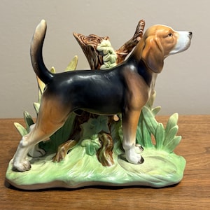 Könnte beinhalten: Eine Keramikfigur eines Beagle-Hundes, der im Profil steht. Der Hund hat ein schwarz-braun-weißes Fell und trägt den Schwanz aufrecht. Er steht vor einem braunen Baumstumpf und hellgrünem Blattwerk auf einem modellierten Sockel.