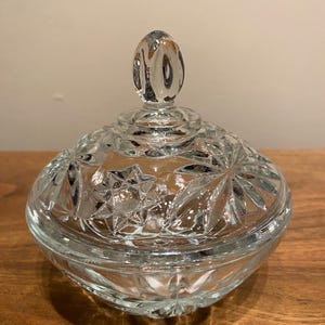 Può includere: Una bomboniera in vetro tagliato trasparente con coperchio. Il piatto è rotondo con un motivo a stella sul coperchio e sui lati. Il coperchio ha una maniglia a forma di goccia. Il piatto è appoggiato su una superficie di legno.