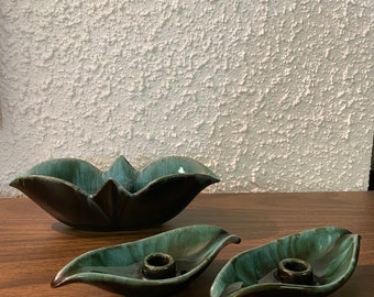 May include: A set of three green and brown ceramic candle holders in the shape of leaves. The holders have a glazed finish and are designed to hold a single candle each.