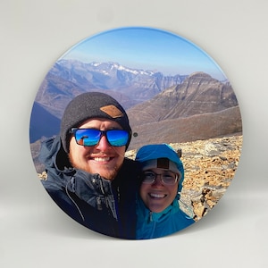 Puede incluir: Una pareja posa para una selfie frente a una cordillera. El hombre lleva una boina negra con un parche marrón, gafas de sol y una chaqueta negra. La mujer lleva una chaqueta azul y gafas.