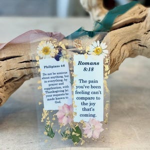May include: Two clear bookmarks with pressed flowers and gold leaf accents. One bookmark features the text "Philipians 4:6" and the other "Romans 8:18". Both bookmarks have a daisy at the top.