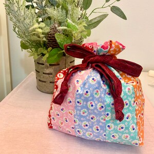May include: A colorful fabric gift bag with a red ribbon and a floral pattern. The bag is tied with a bow and is sitting on a pink surface.