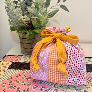 May include: A pink, orange, and white fabric drawstring bag with yellow fabric ties. The bag is made of patchwork fabric with different patterns, including polka dots, gingham, and floral prints.