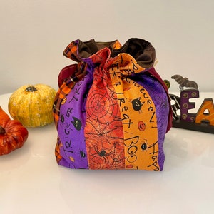 May include: A purple, orange, and black Halloween trick-or-treat bag with a spiderweb design and the words "Trick or Treat", "Happy Halloween", "Boo", and "Treat" printed on it.