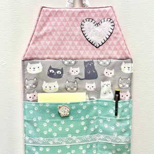 May include: A pink and green fabric wall organizer with a heart-shaped pocket and a pocket with a paw print design. The organizer has a hanging loop and is decorated with a cat print.