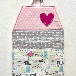Può includere: Un organizzatore da parete in tessuto rosa e grigio con una tasca a forma di cuore e più tasche con stampe a tema gatto. Le tasche sono etichettate con le parole "miao", "purr", "gatto", "gattino" e "gatti".