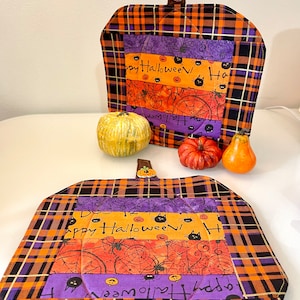 Puede incluir: Dos agarraderas de olla de Halloween de cuadros naranja y negro con un panel de tela morado, naranja y negro con el texto "Happy Halloween!" y gatos negros y arañas.