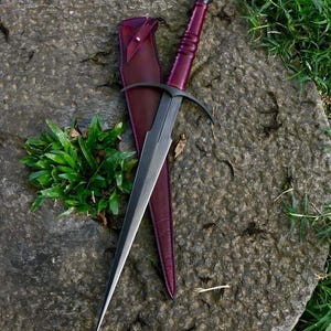 May include: A black dagger with a curved handle and a burgundy leather sheath. The dagger is resting on a gray stone surface with green grass in the background.