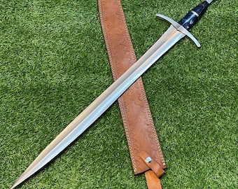 May include: A long, silver sword with a black and silver handle and crossguard rests on a brown leather sheath. The sheath is embossed with a decorative pattern and has a leather strap. The sword and sheath are set against a green background.