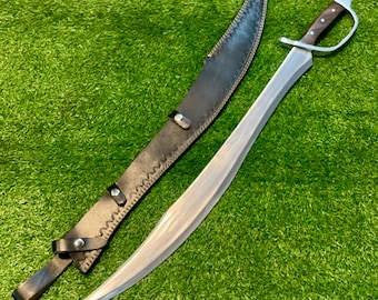 May include: A curved sword with a silver blade and a dark wooden handle, next to a black leather sheath. The sheath is stitched with a light-colored thread and has metal rivets. The sword is on a green surface.