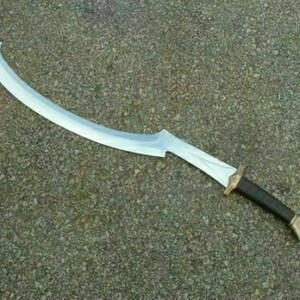 May include: A silver-colored curved sword with a brown leather handle and a gold-colored pommel. The sword is lying on a gray, textured surface.