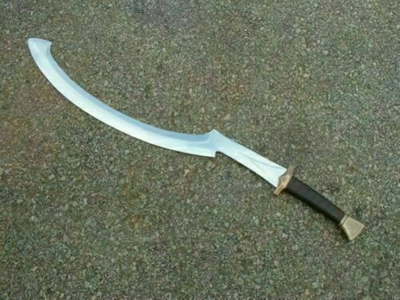 May include: A silver-colored curved sword with a brown leather handle and a gold-colored pommel. The sword is lying on a gray, textured surface.