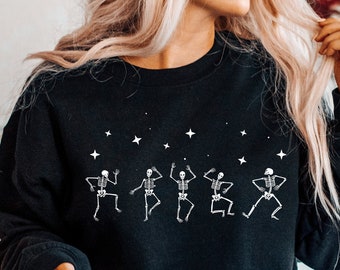 May include: Black sweatshirt with a white graphic of dancing skeletons and stars. The sweatshirt is worn by a person with long blonde hair and is sitting on a white couch.