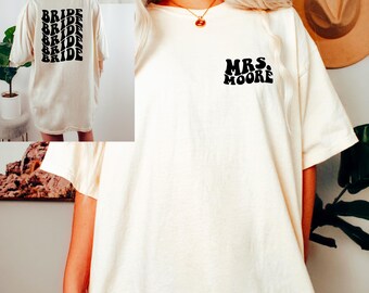May include: A white oversized t-shirt with black text on the front that says "MRS. MOORE" in a retro font. The back of the shirt has the word "BRIDE" repeated five times in a vertical stack in a retro font.