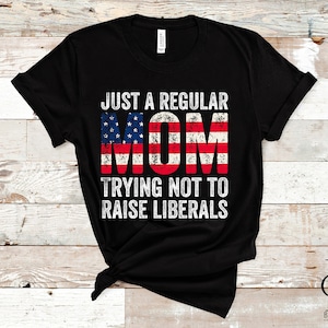 May include: Black t-shirt with the text "JUST A REGULAR MOM TRYING NOT TO RAISE LIBERALS" in white. The word "MOM" is designed with a distressed American flag pattern. The shirt is displayed on a light wood background.