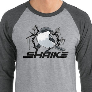 May include: A heather grey and charcoal grey baseball tee shirt with a black and white illustration of a shrike bird perched on a branch. The text "SHRIKE" is printed in black below the bird.