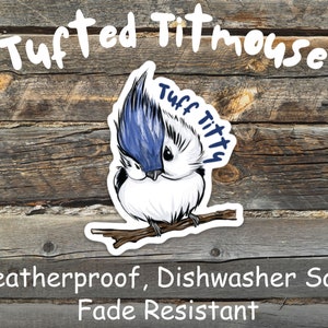 May include: A sticker of a tufted titmouse bird with blue and white feathers. The sticker has the text "Tuff Titty" on it. The sticker is on a wooden background and the text "Weatherproof, Dishwasher Safe Fade Resistant" is below the sticker.