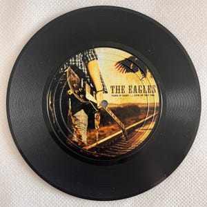 May include: A black vinyl record featuring the band "The Eagles" with the text "Take It Easy... Live in the USA". The record's label shows a person playing a guitar on a railroad track.