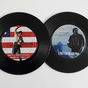 May include: Two black vinyl records featuring Bruce Springsteen album art. One record has the American flag and the text "BRUCE SPRINGSTEEN BORN IN THE U.S.A." The other record has a photo of Springsteen and the text "STREETS OF PHILADELPHIA."