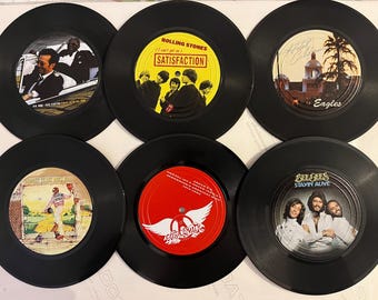 Puede incluir: Seis discos de vinilo negros de 45 RPM con varios artistas y portadas. Las etiquetas incluyen a Eric Clapton, The Rolling Stones Satisfaction, Eagles Hotel California, Elton John, Aerosmith con etiqueta roja y Bee Gees Stayin Alive.