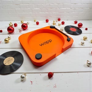 Könnte beinhalten: Ein orangefarbenes Tablett in Form eines Plattenspielers mit dem Wort "wopp" in Schwarz. Zwei schwarze Schallplatten und dekorative Ornamente sind um das Tablett herum verstreut. Das Tablett hat einen schwarzen Tonarm und einen schwarzen Zahnradknopf. Weißer Hintergrund.