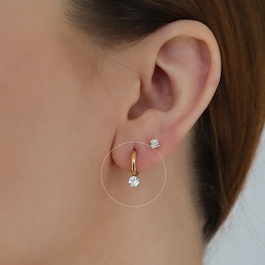 May include: A close-up of a person's ear with two small gold earrings. The earring on the lower lobe has a single clear stone.