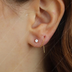 Peut inclure: Une paire de boucles d'oreilles dorées. L'une est un petit clou avec une pierre précieuse transparente, et l'autre est une boucle d'oreille chaîne avec une pierre précieuse transparente.