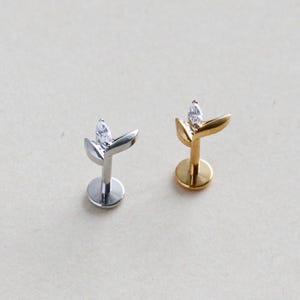 May include: Two flat back labret studs featuring a leaf-shaped design with a single marquise-cut clear gemstone. One stud is finished in a polished silver tone, while the other is finished in a polished gold tone, both with circular bases.