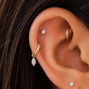 May include: A collection of gold earrings worn in a pierced ear, including a small stud, a hoop with a marquise-cut stone charm, a plain hoop, a second small stud, a thin hoop, and a thick polished gold hoop, all featuring a metallic finish.
