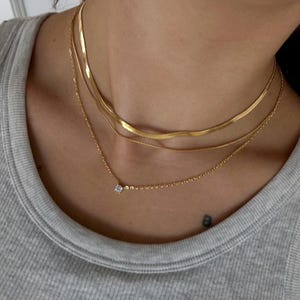 May include: A set of gold necklaces in a layered arrangement. The top layer is a flat, wide chain, the middle is a thinner, rounded chain, and the bottom is a delicate chain with a small, square-cut gemstone pendant. The necklaces are worn over a grey vest top.