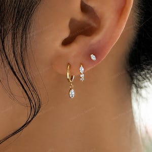 May include: Three gold earrings worn in a single ear. The lowest is a small hoop with a dangling marquise-cut clear stone. The middle is a small hoop with two small clear stones. The highest is a single marquise-cut clear stone stud earring.