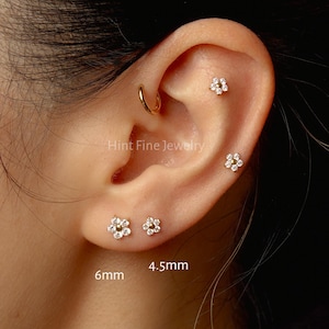 May include: An ear with multiple small, flower-shaped stud earrings. The earrings are set with small, sparkling stones. One earring is a hoop. The text "Hint Fine Jewelry" is visible. The earrings are 6mm and 4.5mm in size.