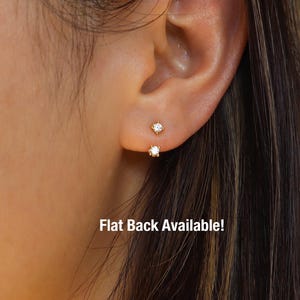 May include: Close-up of an ear with two small, gold-coloured stud earrings. Each earring has a clear, round-cut gemstone. The text "Flat Back Available!" is visible at the bottom of the image.