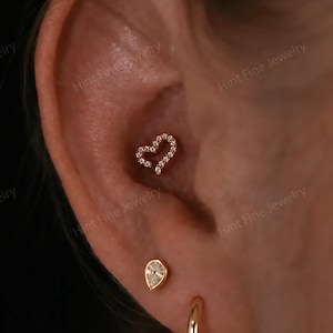 May include: A gold conch piercing featuring a heart-shaped stud encrusted with small clear stones, a teardrop-shaped stud in the lobe, and a plain gold hoop earring positioned below it, all set against a dark background with a watermark.