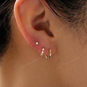 May include: A set of three gold earrings worn in a single earlobe. The arrangement includes one small gold stud with a clear stone, one small gold hoop featuring three tiny clear stones, and one plain, smooth gold hoop earring.