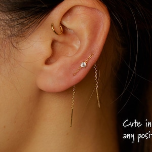 May include: Close-up of a gold ear piercing with a small, clear gemstone and a delicate chain. A gold hoop earring is also visible. The text "Cute in any position" is written in cursive.