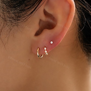 May include: A set of three gold-toned earrings worn in the earlobe. The set includes one plain polished gold hoop, one small gold hoop featuring three tiny clear stones, and one single stud earring with a small clear stone at the top.