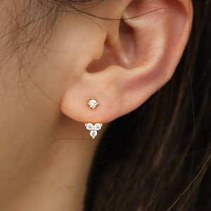 May include: A pair of gold earrings with small clear cubic zirconia stones. The earrings are a single stone on the top and a cluster of three stones dangling below.