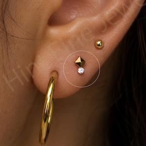 May include: An ear with gold earrings. A large gold hoop earring is in the lobe, with a small gold stud and a gold ball stud above it. A small gold earring with a diamond-like accent is also visible.