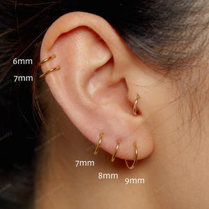 May include: A set of thin gold hoop earrings worn in multiple ear piercings. The hoops are labelled with sizes 6mm, 7mm, 7mm, 8mm, and 9mm. These small, circular metal rings are positioned along the cartilage and the earlobe in a series.