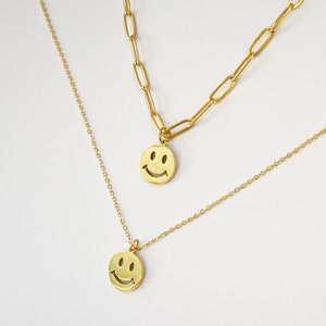 May include: Two-layer gold-colored necklaces. The top layer features a chain with elongated links, while the bottom layer has a delicate beaded chain. Each necklace has a round pendant with a smiling face.