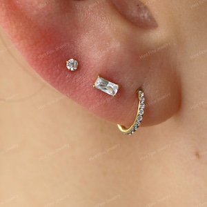May include: A set of three gold-toned earrings worn in an earlobe. The set includes a small round stud with a clear stone, a rectangular baguette-cut stud with a clear stone, and a small gold hoop earring encrusted with tiny clear stones.