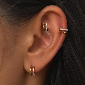 Peut inclure: Boucles d'oreilles créoles et manchette d'oreille dorées avec de petites pierres précieuses transparentes. Les boucles d'oreilles sont à double anneau et la manchette a deux rangées de pierres. Bijoux sur une oreille.