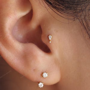 May include: A gold tragus earring featuring a small pear-shaped clear stone with a tiny round stone dangling below. The ear also displays two gold lobe earrings with round clear stones, a gold hoop, and a gold eternity band with clear stones.